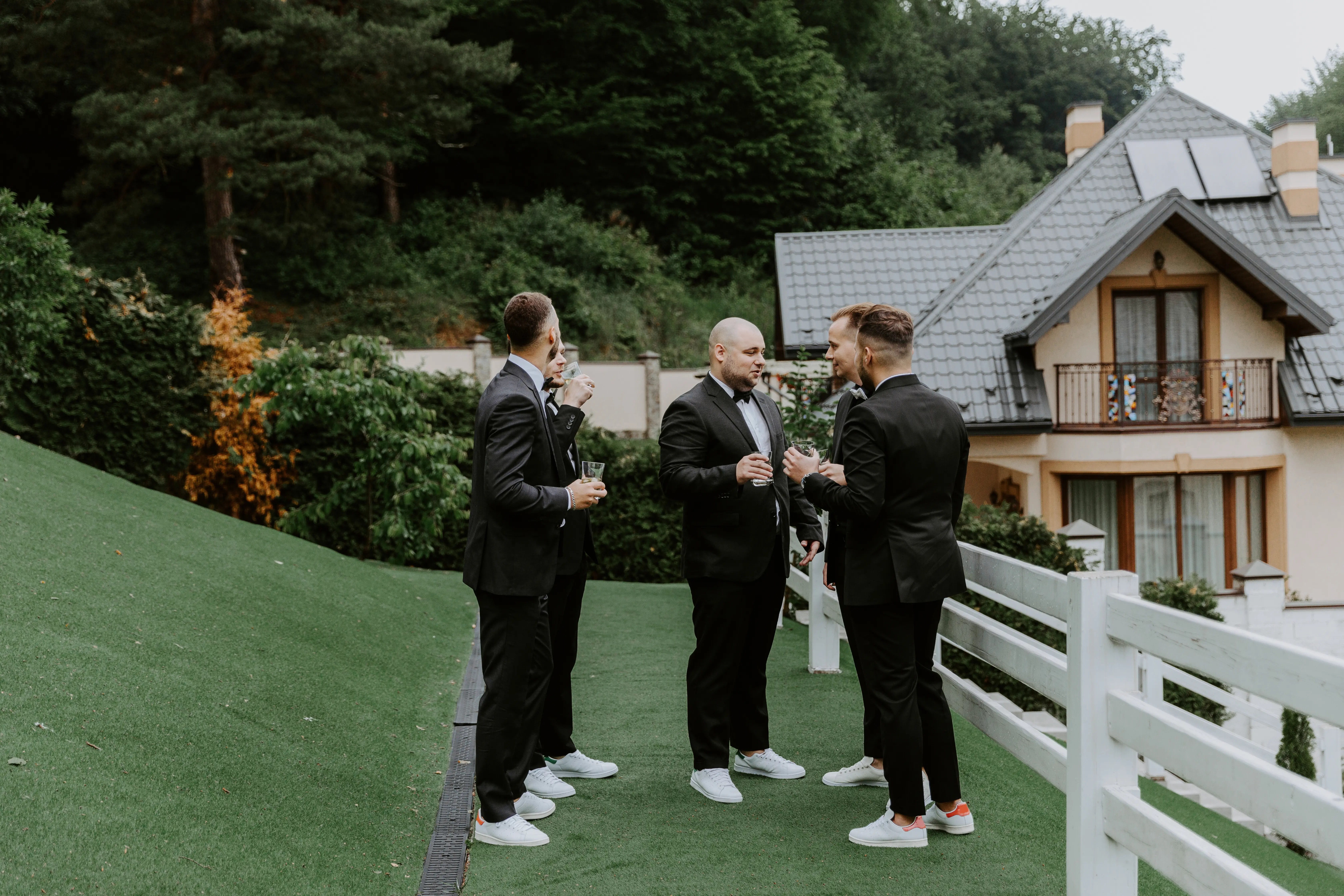 The groom and his groomsmen friends, dressed in stylish suits, drinking whiskey outdoors.